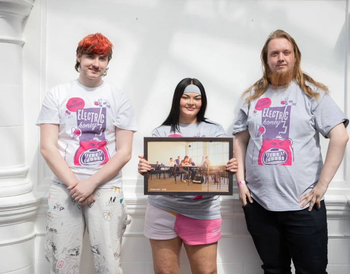 Three students wearing Electric Honey t-shirts standing in front of a white wall, holding a framed classroom photo from 2008. Three students wearing Electric Honey t-shirts standing in front of a white wall, holding a framed classroom photo from 2008.